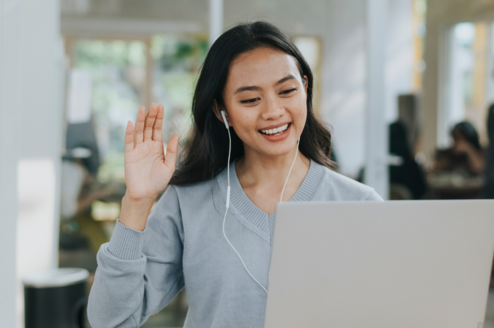 A person with brunette hair and headphones on waves at a computer camera